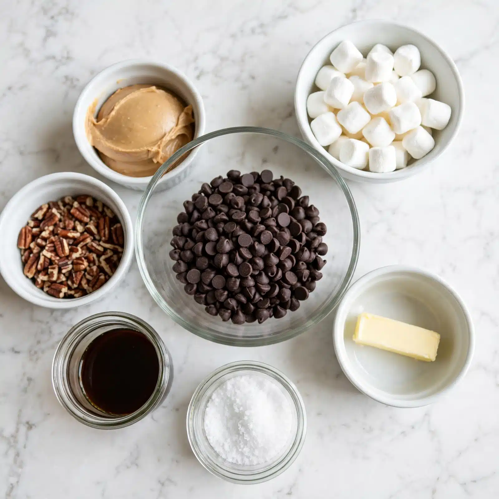 A top view of seven small white bowls and a clear glass bowl on a white marbled surface. The bowls hold different ingredients: one is full of dark chocolate chips in the center with a smooth glass texture; to the upper right, a white bowl with small white marshmallows; to the lower right, a white bowl with a small stick of pale yellow butter; to the left, a white bowl filled with chopped brown pecans; above it, a white bowl with beige creamy peanut butter; below the pecans, a small glass container with dark brown vanilla extract; next to it, a small glass container with coarse salt. photo taken with an iphone --ar 4:5 --v 7